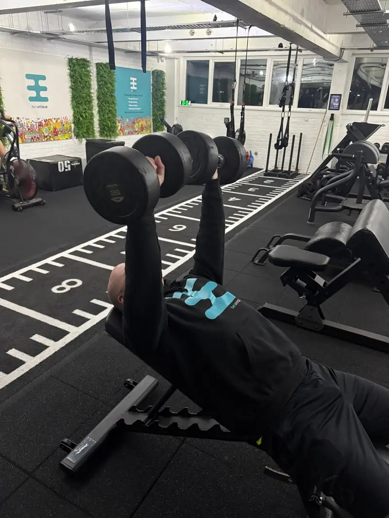 A member pressing dumbbells on a bench with a coach close by
